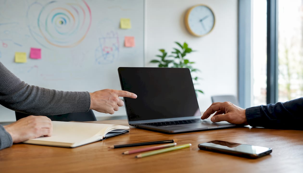 Tijdens een IT-overleg wijzen twee mensen tegelijk naar de laptop en de notities op tafel.